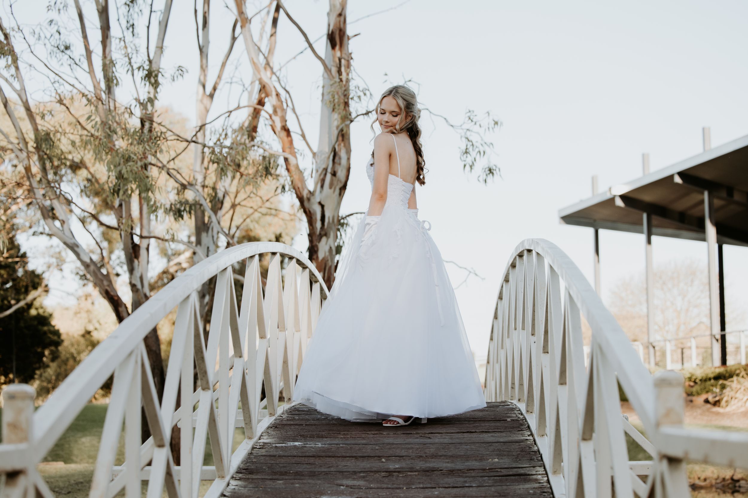 girl in white dress walks along a wooden boardwalk surrounded by eucalyptus trees at sunset.
