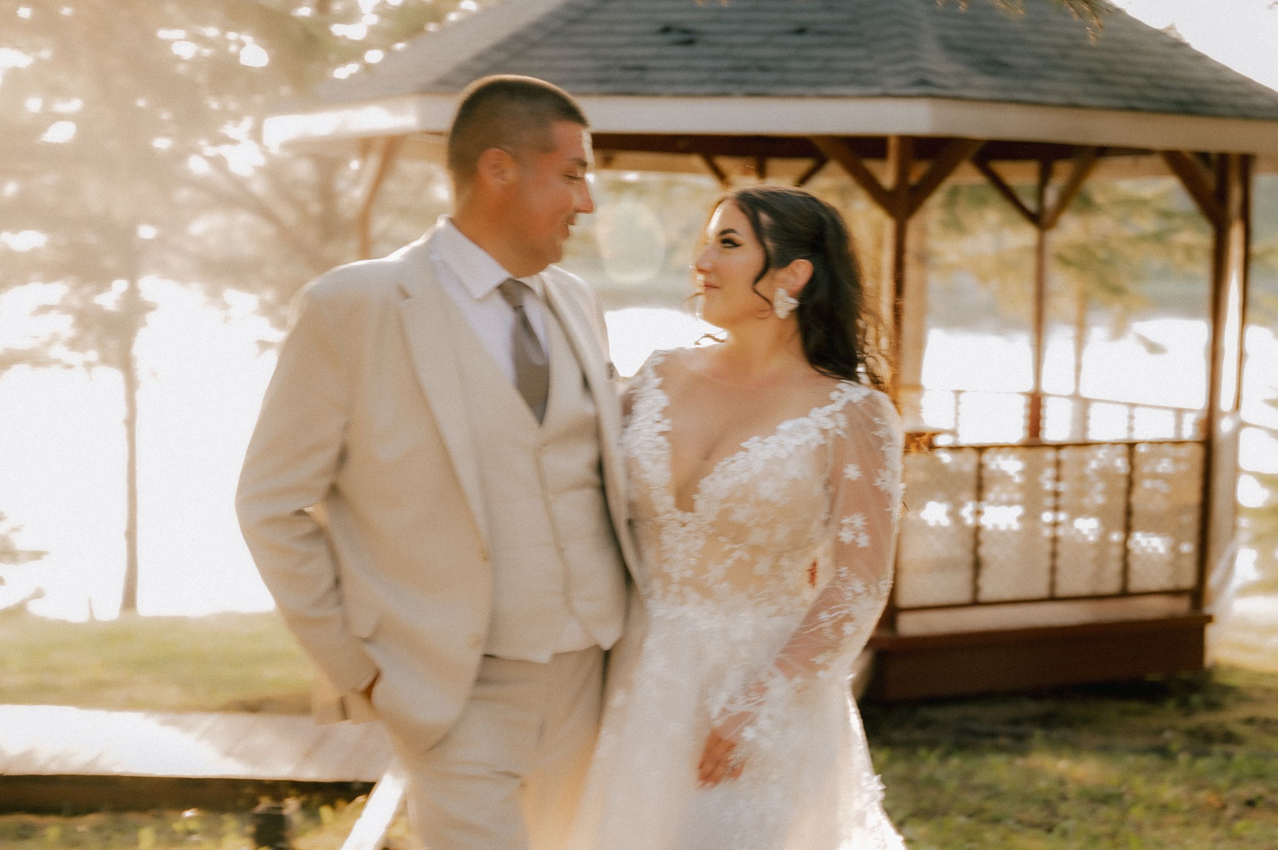 A couple in wedding attire share a romantic moment near a gazebo during sunset, with soft golden light surroundings.