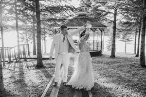A white lace wedding dress flows in the sunlight among evergreen trees and a rustic gazebo background.