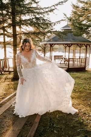 An elegant wedding dress with lace details twirls in golden afternoon light among pine trees.