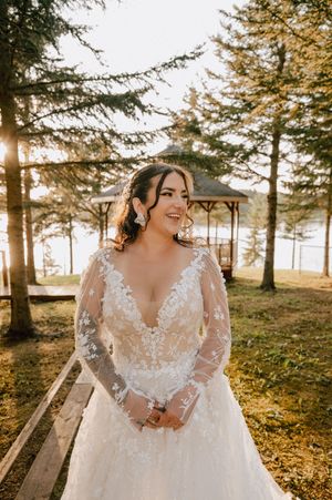 A flowing white wedding gown with long lace sleeves captures sunlight filtering through forest trees.