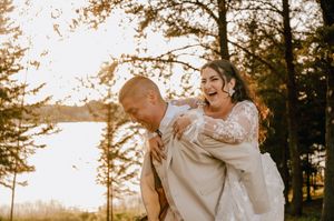 A playful wedding moment with bride riding piggyback on groom's back in a sun-drenched forest setting.
