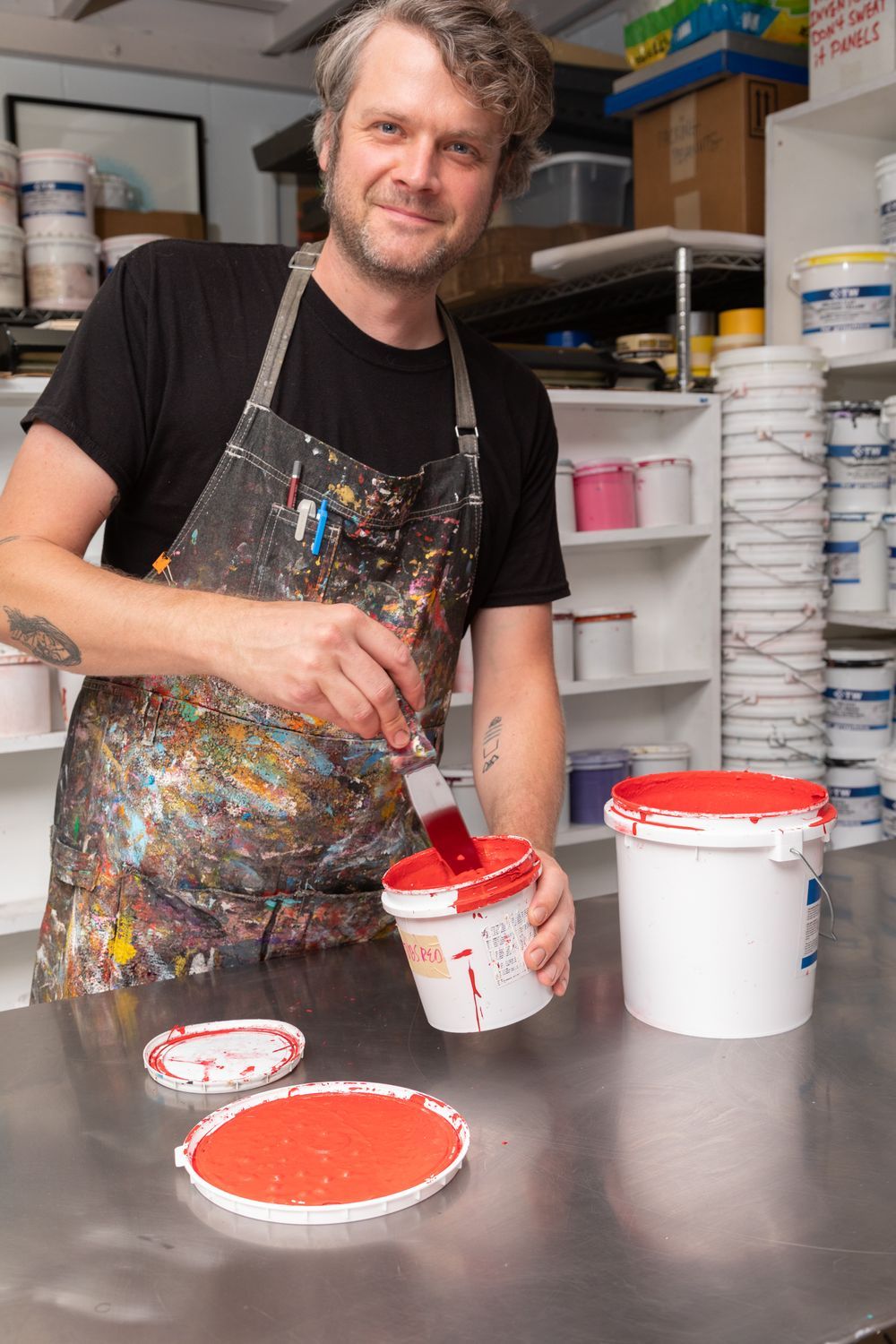 Artist in his studio mixing a bucket of red paint, while wearing an apron covered in paint colors.