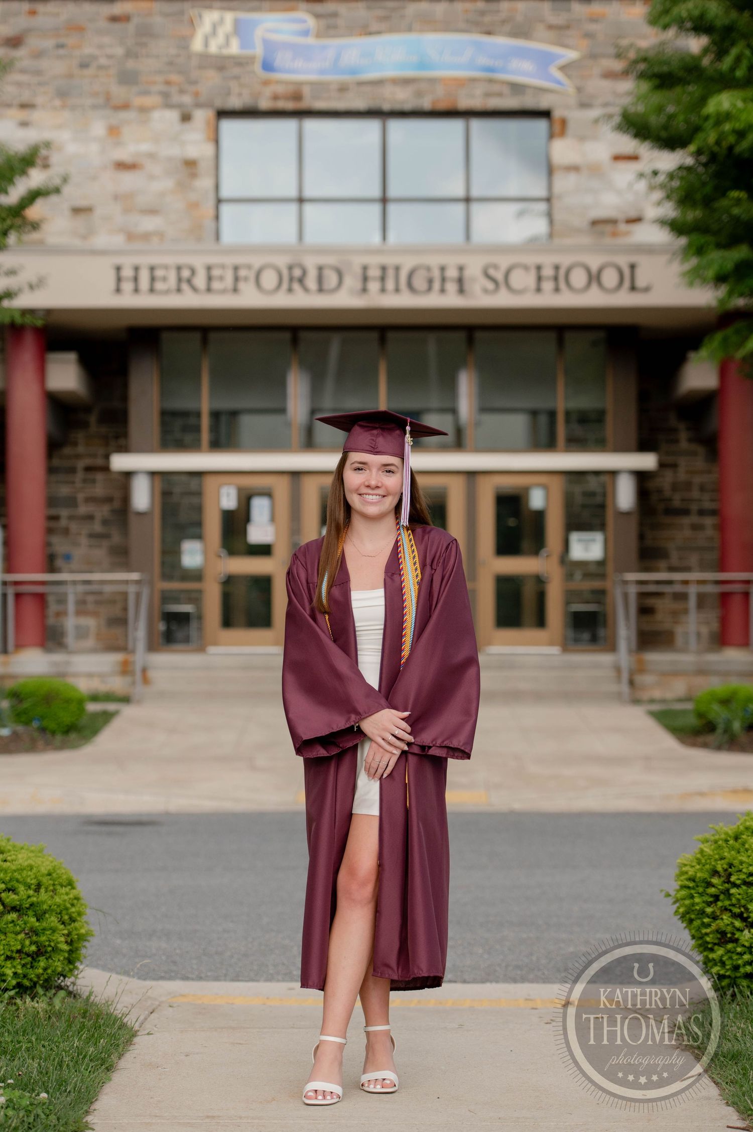 Graduate in maroon cap and gown poses in front of Hereford High School entrance on graduation day.