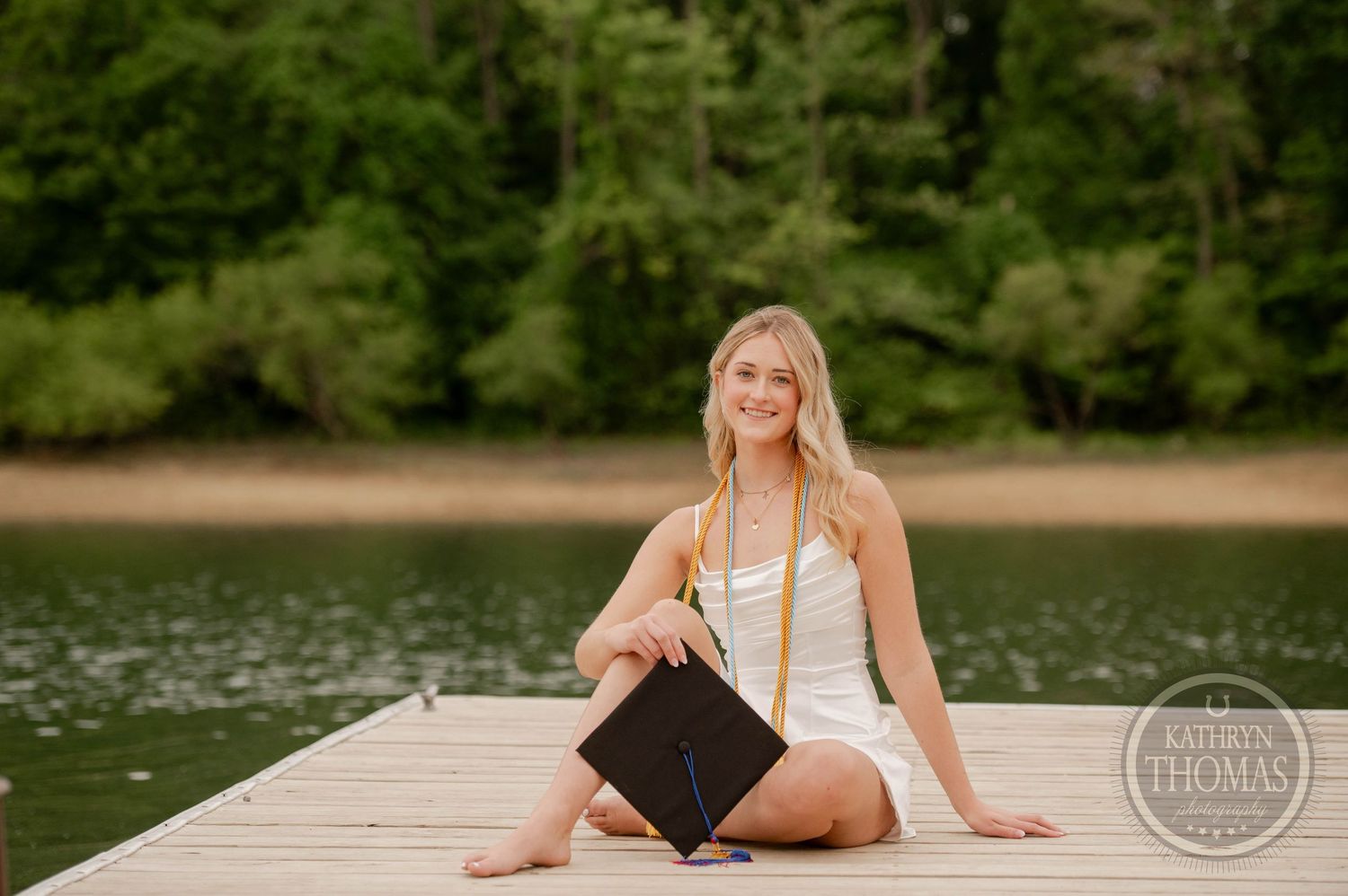 Summer senior portrait session of a student sitting on a wooden dock by a serene lake surrounded by green forest.