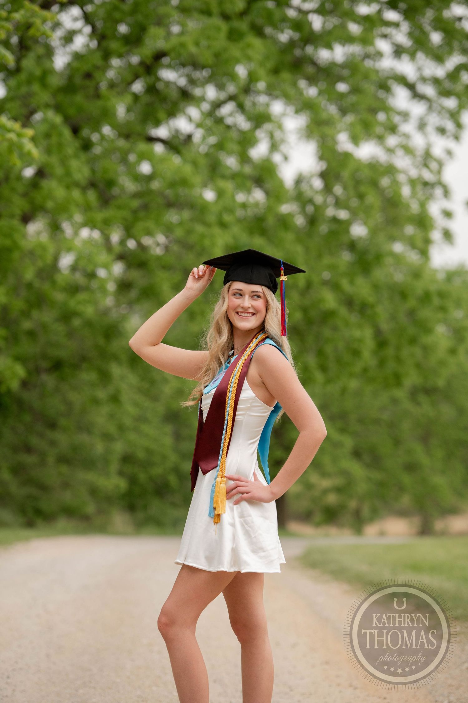 Graduate poses playfully in white dress and cap with stole and honor cords against lush green outdoor backdrop.