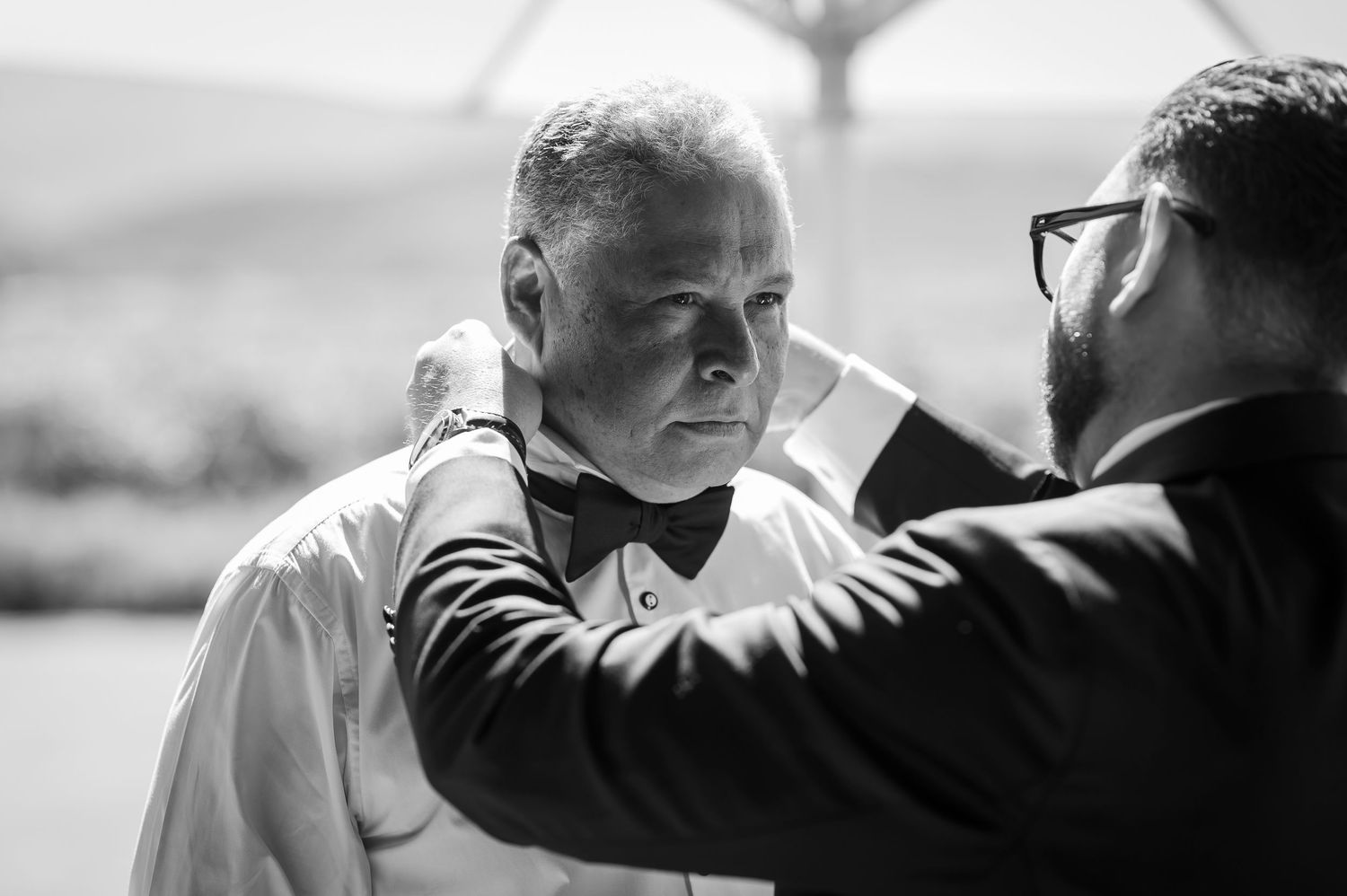 A black and white photo captures an intimate moment of adjusting a bow tie before a wedding ceremony.
