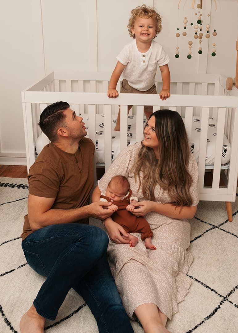 Toddler climbs on white crib while parents sit on patterned rug with newborn in cozy nursery setting at a family lifestyle session