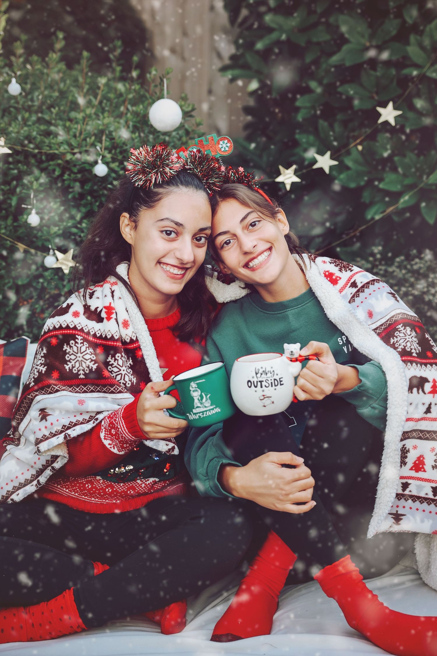 Two friends in festive Christmas sweaters holding holiday mugs and smiling while surrounded by decorative stars and ornaments.