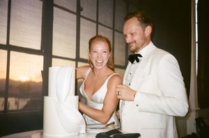 A couple in white formal attire laughs and embraces during what appears to be a wedding celebration at sunset.