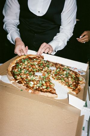 Person in formal attire serving a pizza topped with fresh herbs and toppings from a cardboard delivery box.