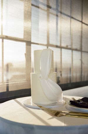 Modern white wedding cake with geometric bow design sits elegantly on table in natural sunlight.