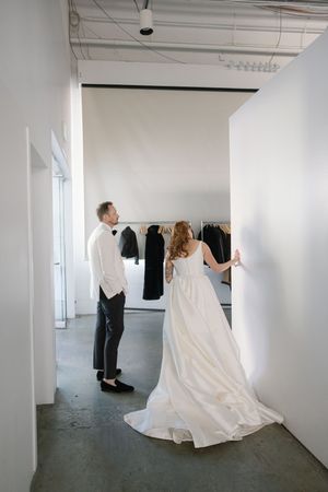 A couple in wedding attire examines clothing on hangers in a minimalist white room.