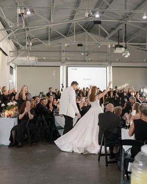 A wedding reception in an industrial-style venue with guests seated at tables applauding.
