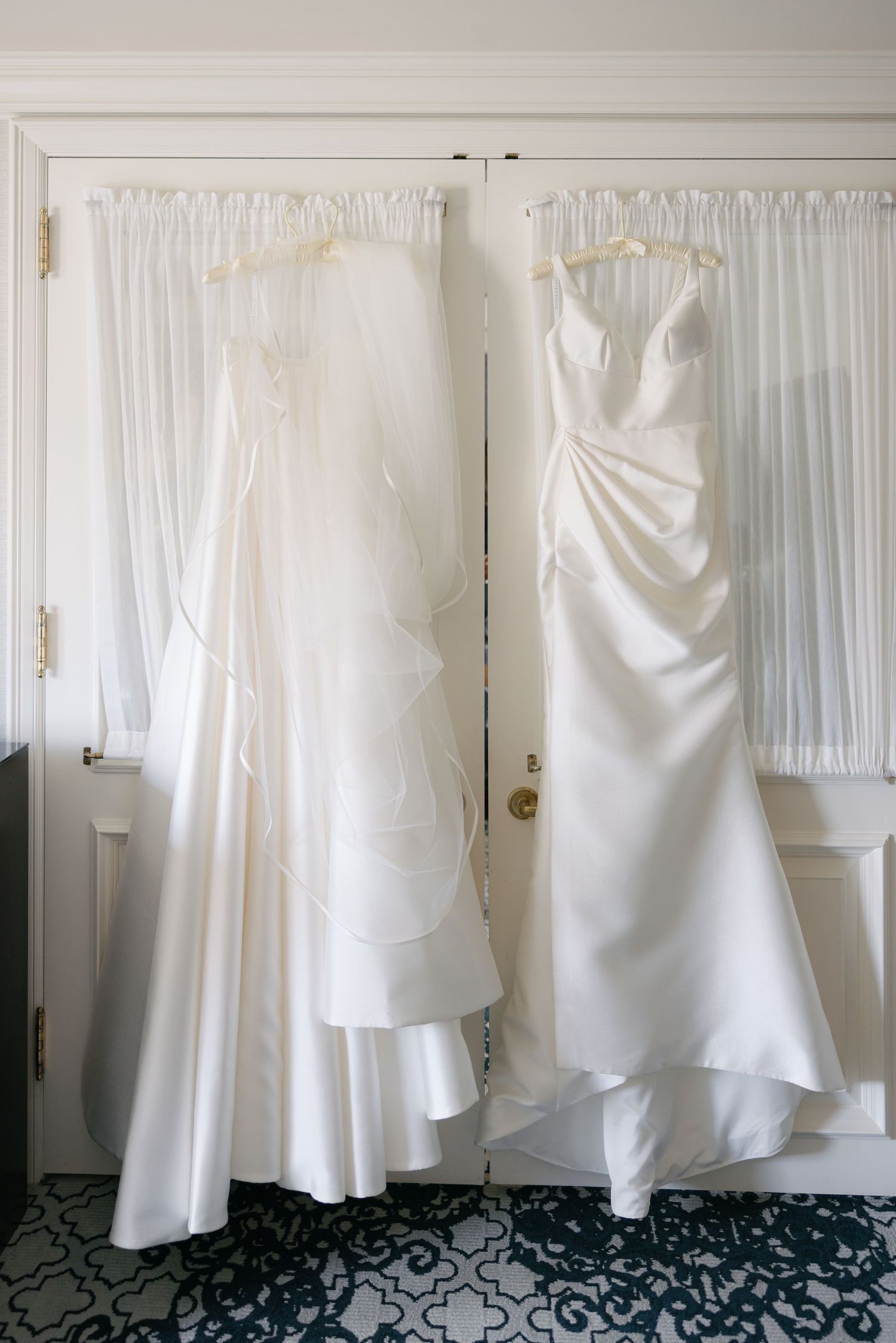 Two elegant white wedding dresses hanging on display against a curtained wall, showcasing different silhouettes and styles.