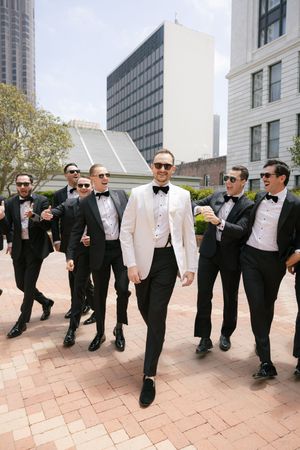 Groomsmen in tuxedos strike playful poses together on outdoor patio.