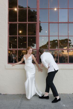 A couple in wedding attire poses playfully by a large window at sunset.