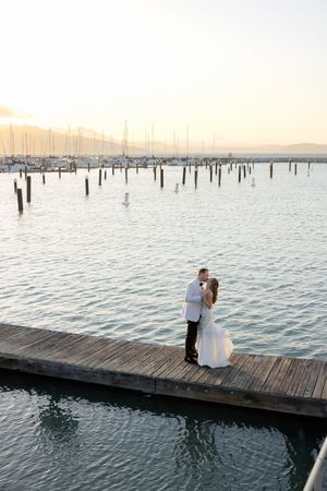 A couple embraces on a wooden dock at sunset with sailboats and harbor posts visible in the background.