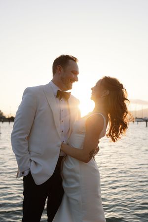 A couple shares an intimate moment at sunset by the waterfront in formal wedding attire.
