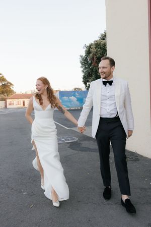Wedding couple runs and dances together on a concrete area beside a white building.