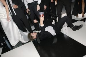 Party guest in formal attire lays on dance floor taking photo while others stand around during celebration event.
