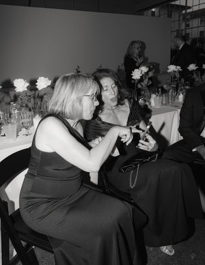Two guests in formal black dresses engage in conversation at an elegant dinner table with floral centerpieces.
