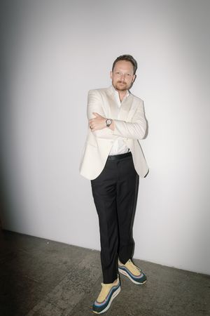 A person in a white blazer and black pants poses casually against a light grey wall.