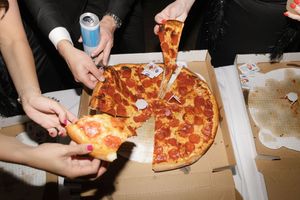 Hands reaching for slices of pepperoni pizza from an open cardboard delivery box on a table.