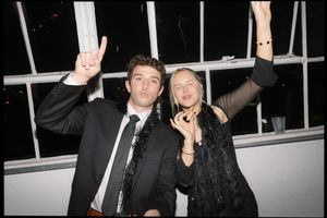 Two party guests throw their hands up in celebration while posing for a nighttime photo at an indoor event.