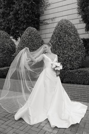 A bride in an elegant white gown poses with flowing veil against manicured hedges and brick wall backdrop.