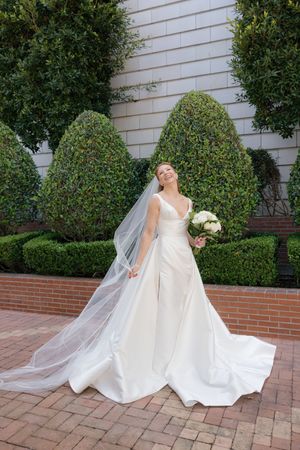 A bride in an elegant white wedding gown and veil poses gracefully on a brick patio surrounded by manicured shrubs.