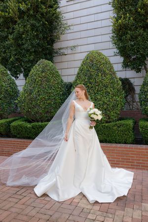 A bride in a flowing white dress with veil holds a bouquet while posing on red brick pavers next to topiary bushes.