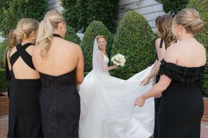 Bridesmaids in black dresses helping adjust bride's white wedding gown outdoors.