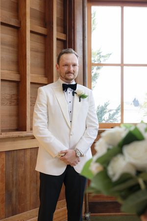 Groom in white dinner jacket and black bow tie poses by window in wooden interior.