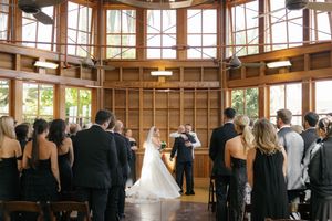 A wedding ceremony takes place in a sunlit wooden room with high windows and standing guests.