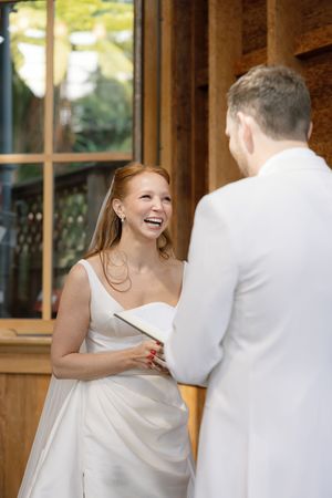 A bride in a white wedding dress shares a joyful laugh with the groom  warmly lit wooden room. Sunnyside conservatory in San Francisco