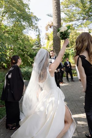 A bride in a white dress raises her bouquet while celebrating outdoors with wedding party members.