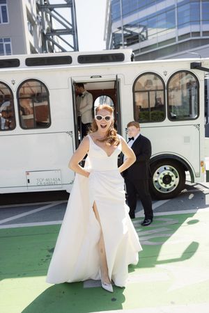 Bride in white dress poses playfully near vintage bus on sunny day.