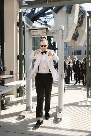 A groom in a white dinner jacket and bow tie poses confidently in an outdoor walkway.