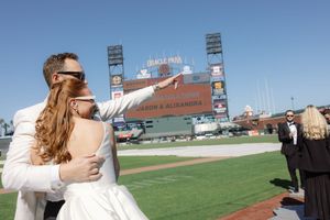 A wedding photo taken at a baseball stadium with the iconic stadium architecture visible in the background.