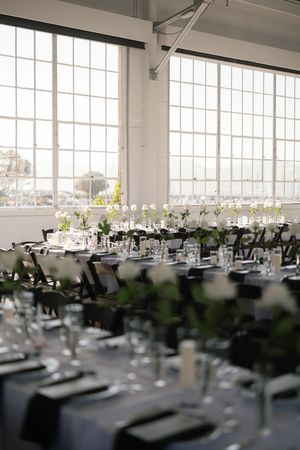A light-filled modern reception venue with rows of elegantly set tables and large industrial windows.