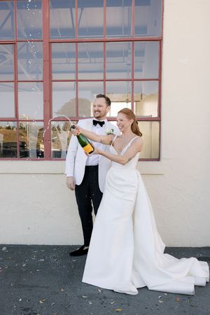 Couple in wedding attire popping champagne outside a building with red-framed windows.