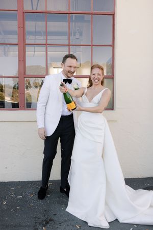 A couple celebrates with champagne outside a building, dressed in elegant white wedding attire.