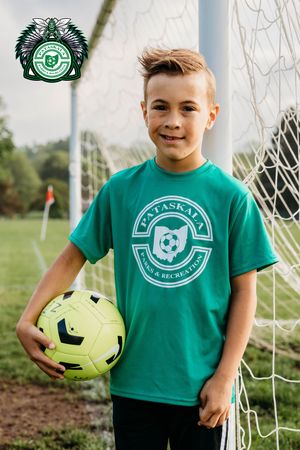 Young soccer player wearing green Savannah t-shirt poses with yellow soccer ball by goal post on grass field.
