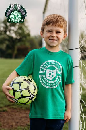 Young soccer player in a green club t-shirt poses with a soccer ball next to a goal post.