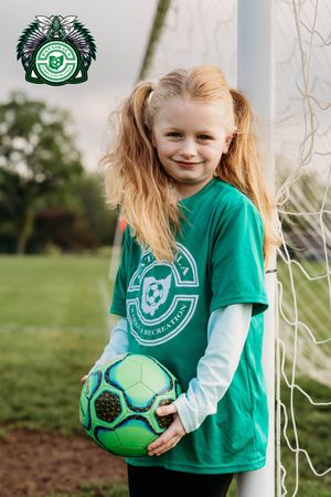 Young soccer player in green long-sleeved uniform shirt holds green soccer ball while standing by goal post on grass field.