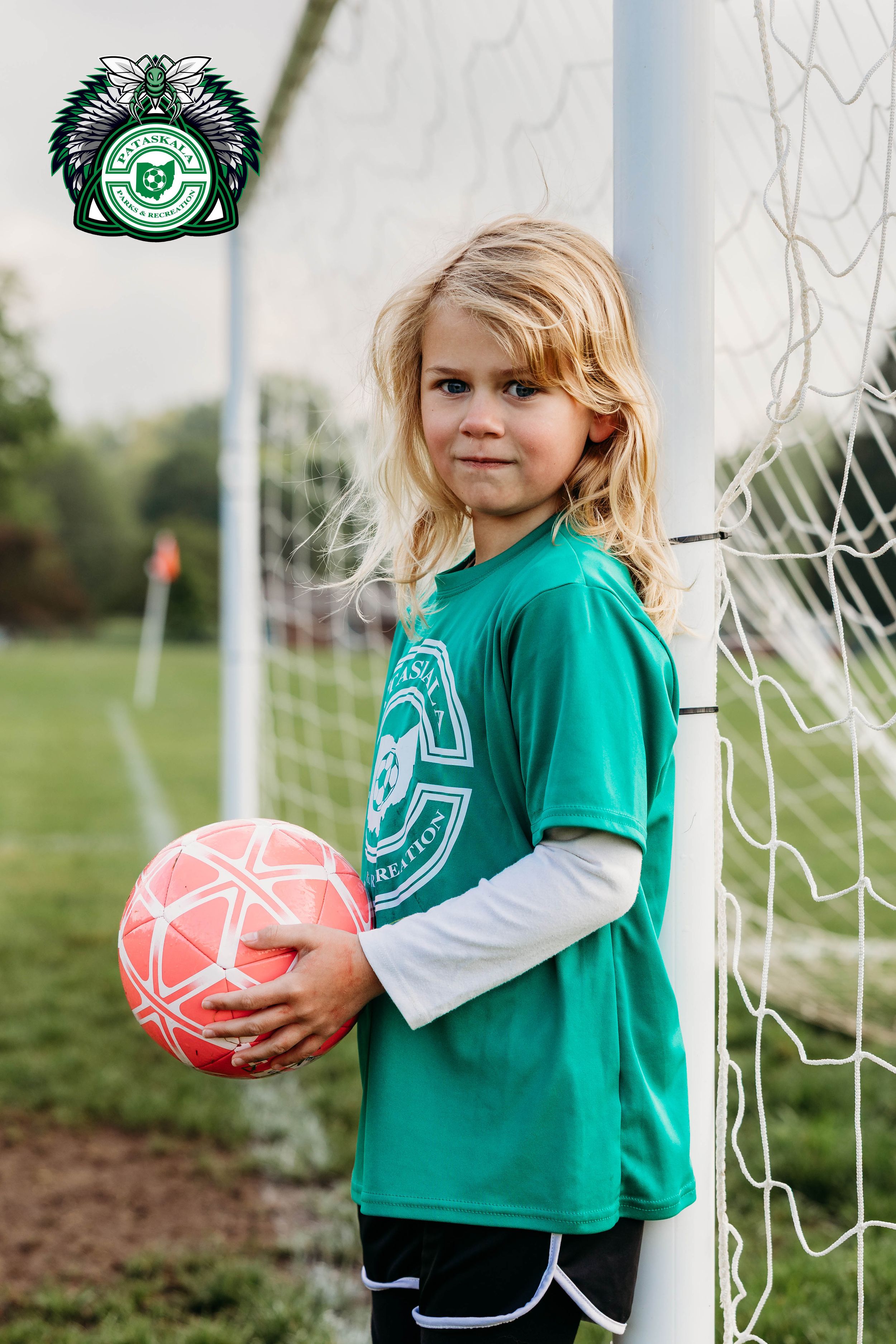 Young soccer player in green jersey poses with pink ball next to goal post.