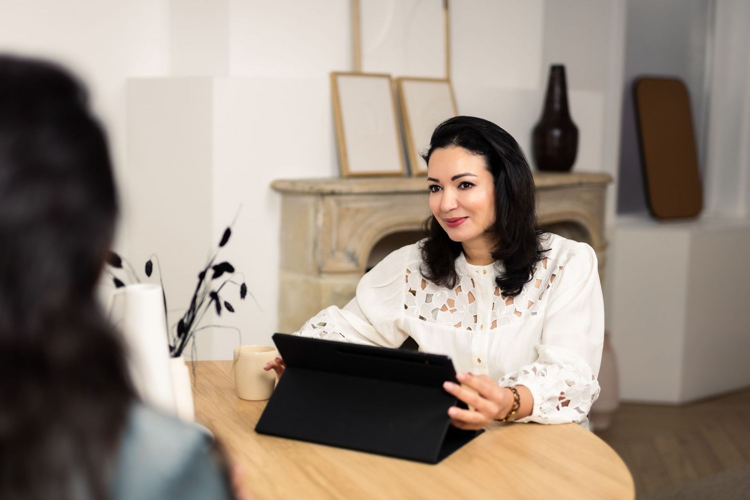 Professional wearing white blouse smiles while working on digital tablet at wooden desk in modern home office setting.