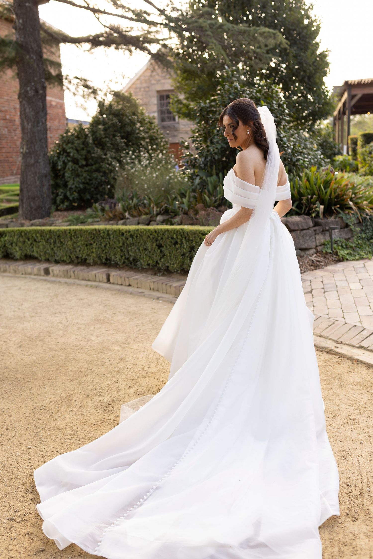 Couple walking along garden path as bride's elegant white wedding dress train flows behind them at sunset.