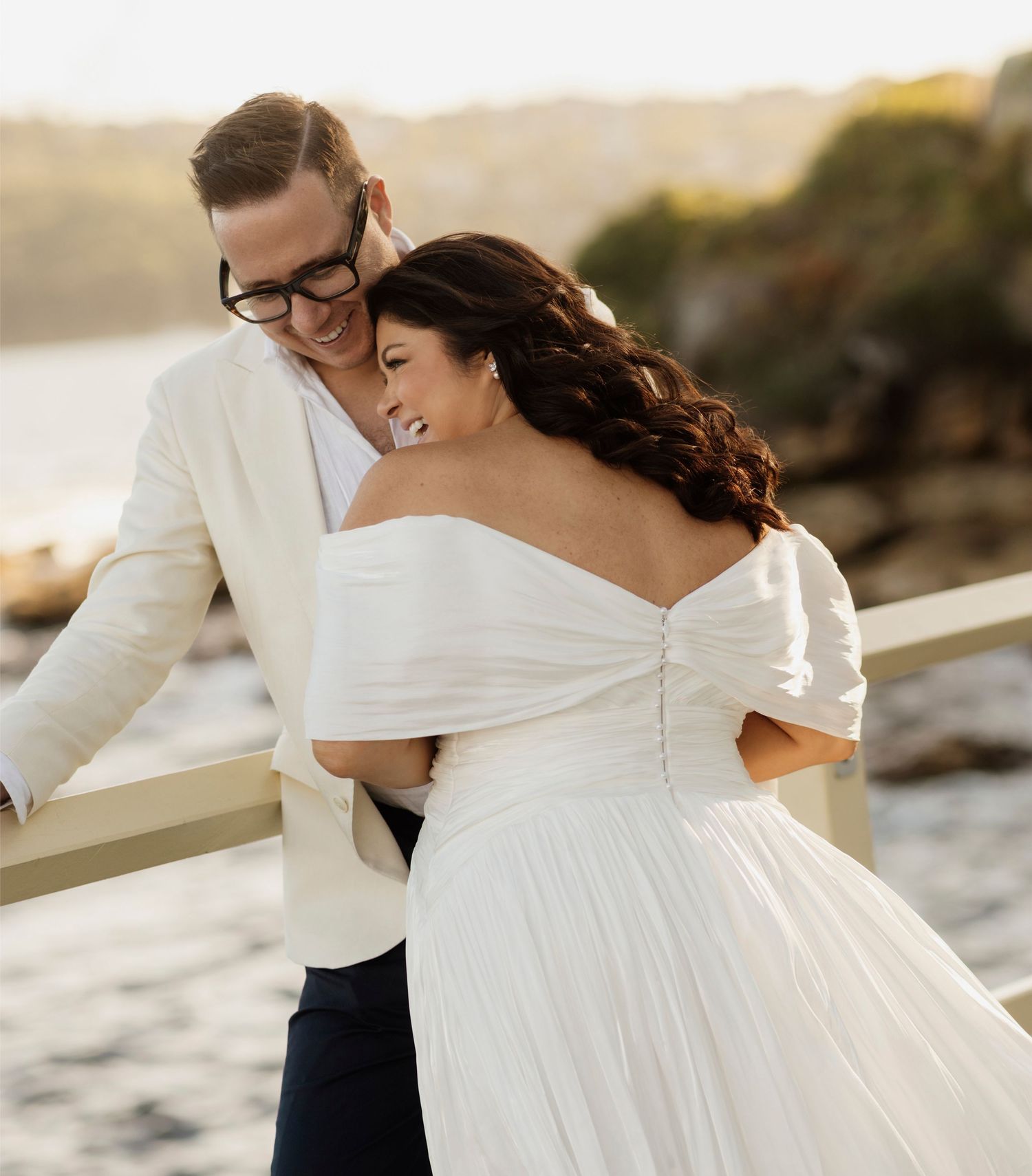 A couple shares an intimate moment at sunset on a rocky coastline in formal wedding attire.